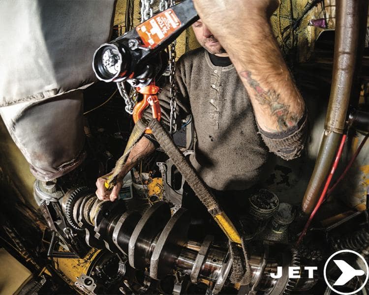 Mécanicien tatoué utilisant un palan industriel JET Tools pour l'entretien d'une machine dans un atelier bien rempli.