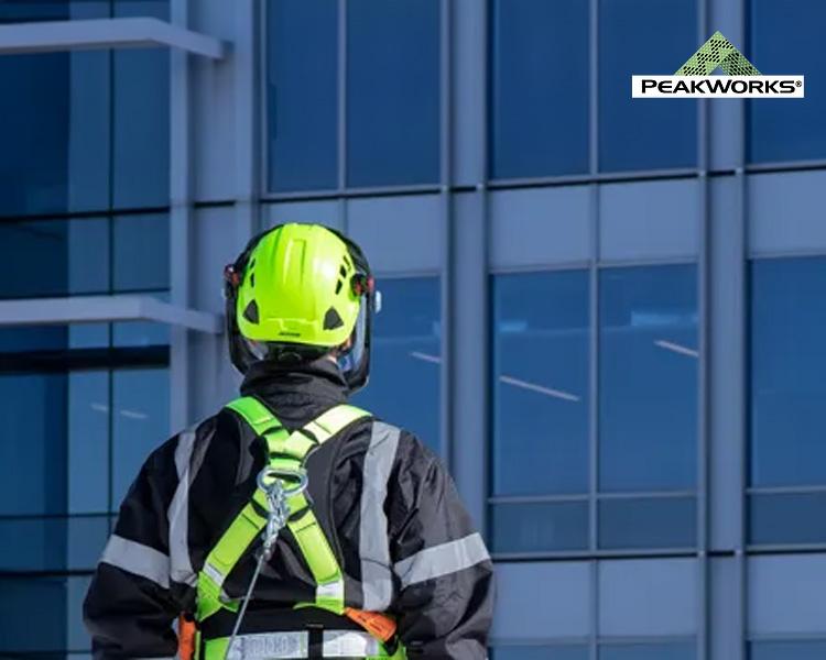 Un travailleur en équipement de sécurité, portant un casque et un harnais, observe un grand bâtiment en verre de l'extérieur, avec le logo Peakworks visible dans le coin.
