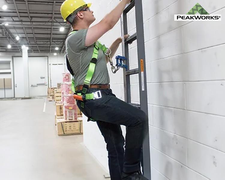 Un ouvrier de construction avec un casque jaune et un harnais vert installe un équipement sur un mur dans un entrepôt, illustrant la sécurité et la précision au travail.