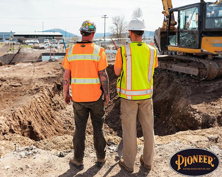 Deux travailleurs de la construction en gilets haute visibilité et casques se tiennent près d'un chantier d'excavation avec un bulldozer en arrière-plan, discutant du projet.