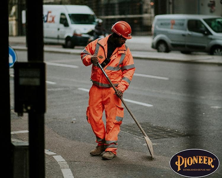 Un travailleur de la construction en tenue réfléchissante orange utilisant une houe dans une rue de la ville, avec des véhicules en arrière-plan et le logo Pioneer visible.